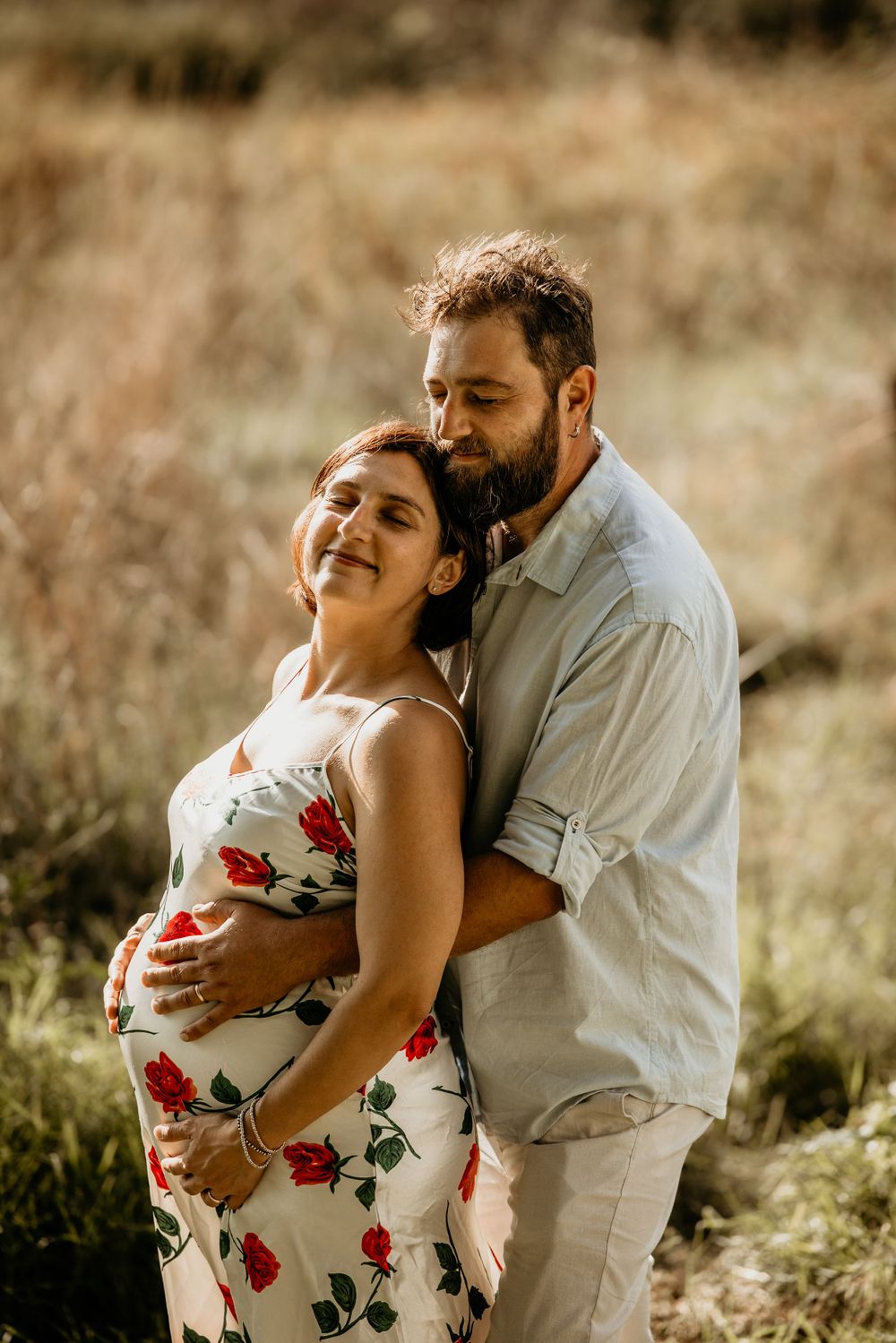 Man embracing pregnant woman in floral dress from behind in golden dry grass field.