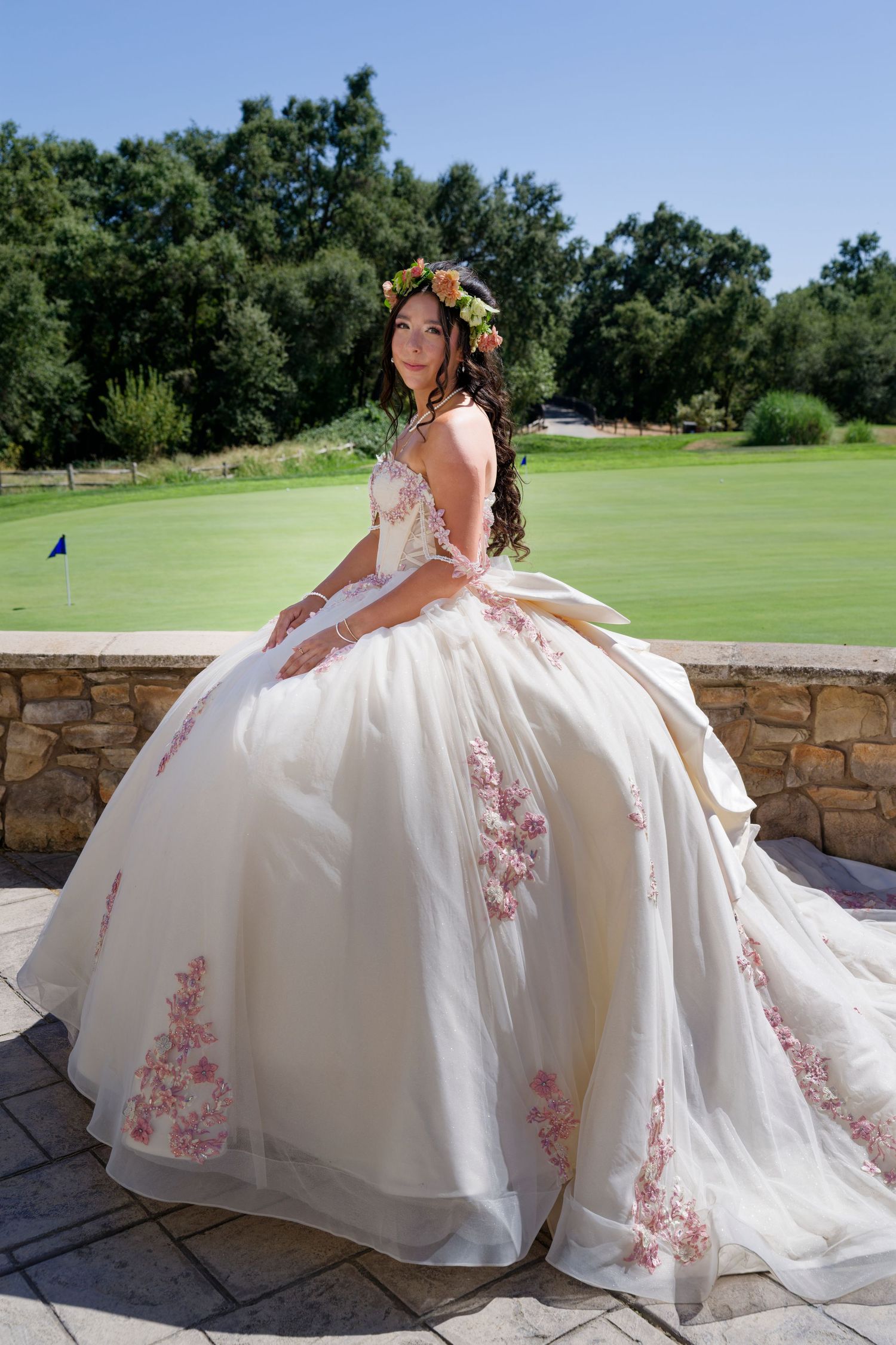 Young woman in elegant white quinceañera ball gown with pink floral embroidery and flower crown poses outdoors.