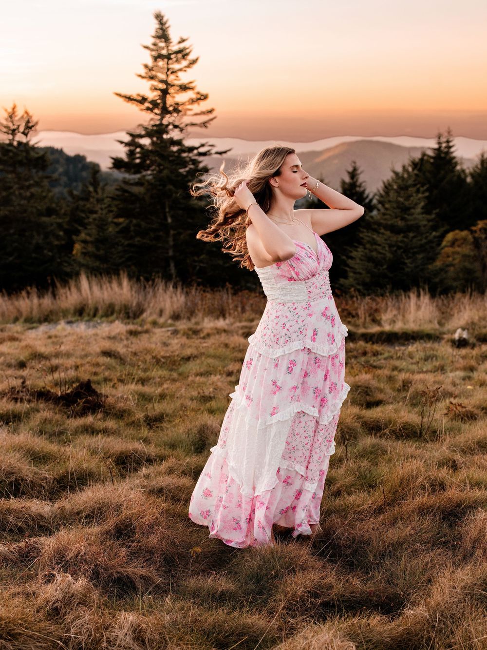 A woman in a pink floral maxi dress poses gracefully on a hillside at sunset with misty mountains in the background.
