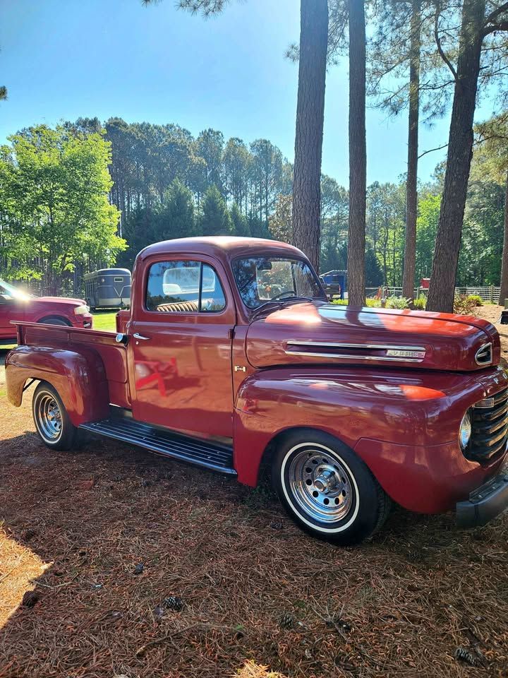 Restored vintage red Ford F-1 pickup truck parked among tall pine trees on a sunny day.
