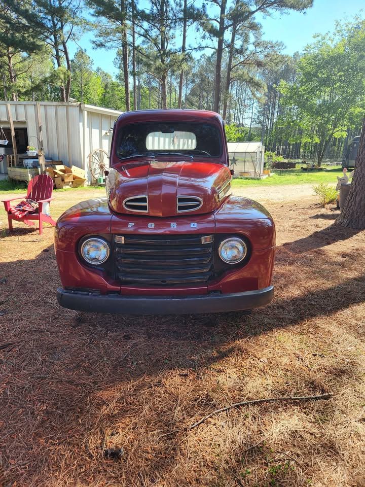 Restored classic red Ford pickup truck from the 1950s parked on a pine needle covered driveway surrounded by tall trees.