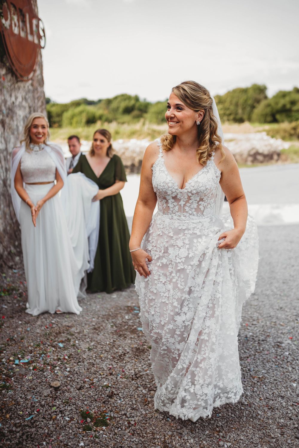 Smiling bride in a lace wedding gown walks along a gravel path while bridesmaids and guests stand behind her.