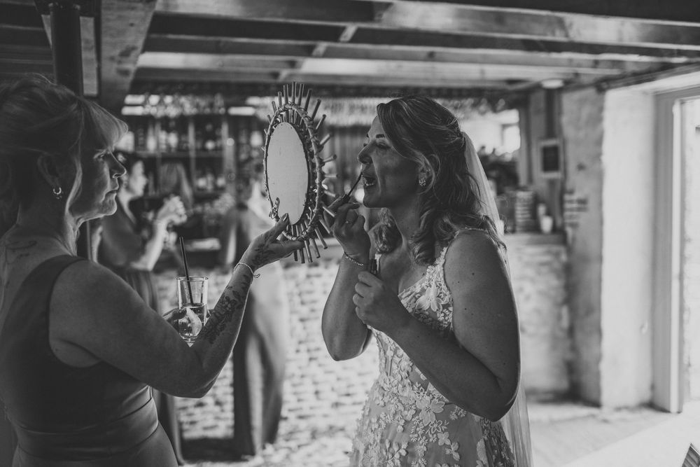 A bride in a lace wedding dress smiles at herself in a sunburst mirror held by a woman while getting ready for her wedding.