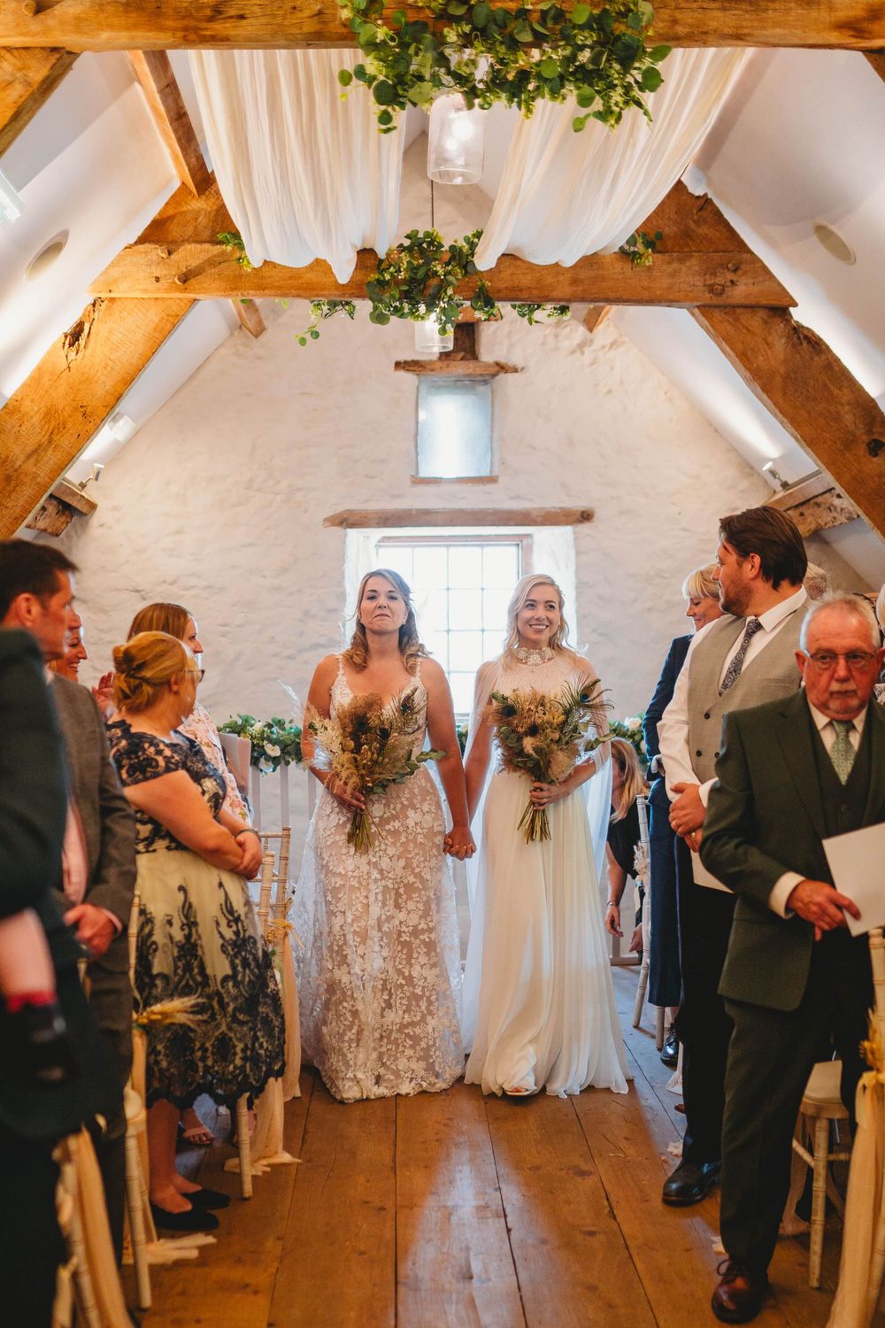 Two brides in lace and white gowns hold hands and bouquets walking down the aisle of a rustic barn wedding venue as guests watch.