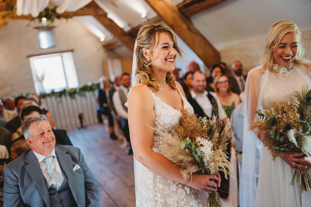 Smiling bridesmaids in white lace dresses hold wildflower bouquets while wedding guests watch the barn ceremony.