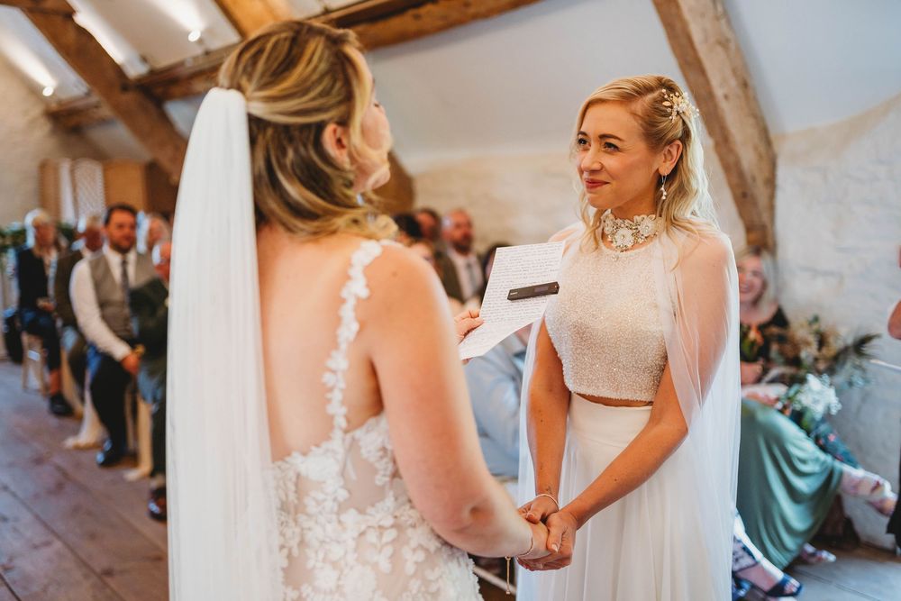 Two brides in white lace wedding gowns and veils hold hands while exchanging vows during a rustic indoor ceremony with guests watching.