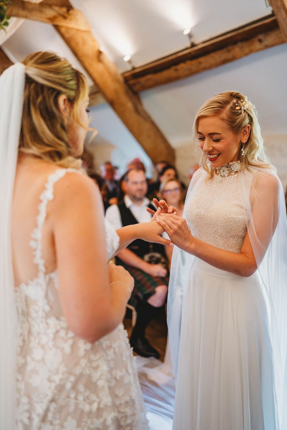Smiling bride in a white lace gown and veil exchanges rings with her partner during a rustic wedding ceremony as guests watch.