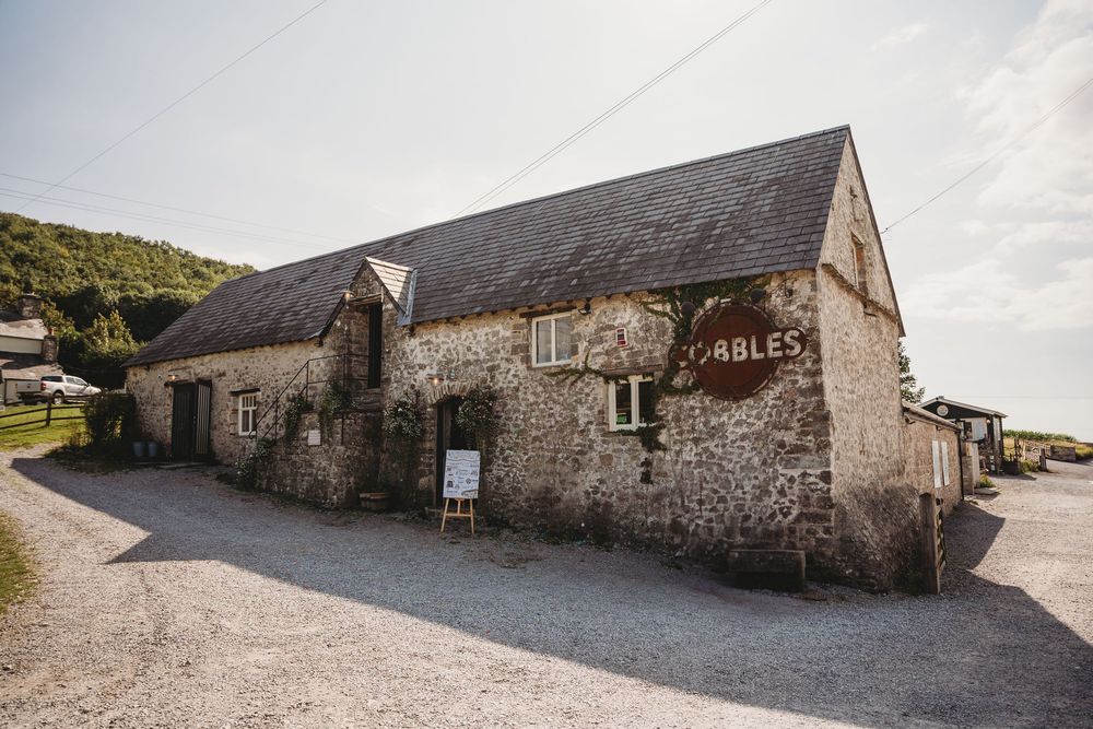 Rustic stone barn building with a slate roof, ivy-covered walls and a round sign, set beside a gravel courtyard with hills behind.