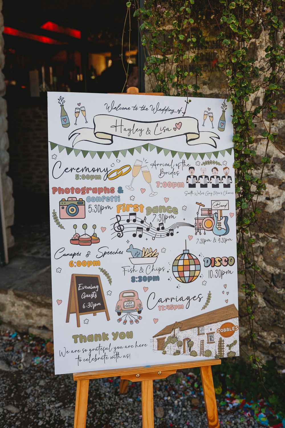 Illustrated wedding day timeline welcome sign for Hayley and Lisa on a wooden easel beside an ivy-covered stone wall.