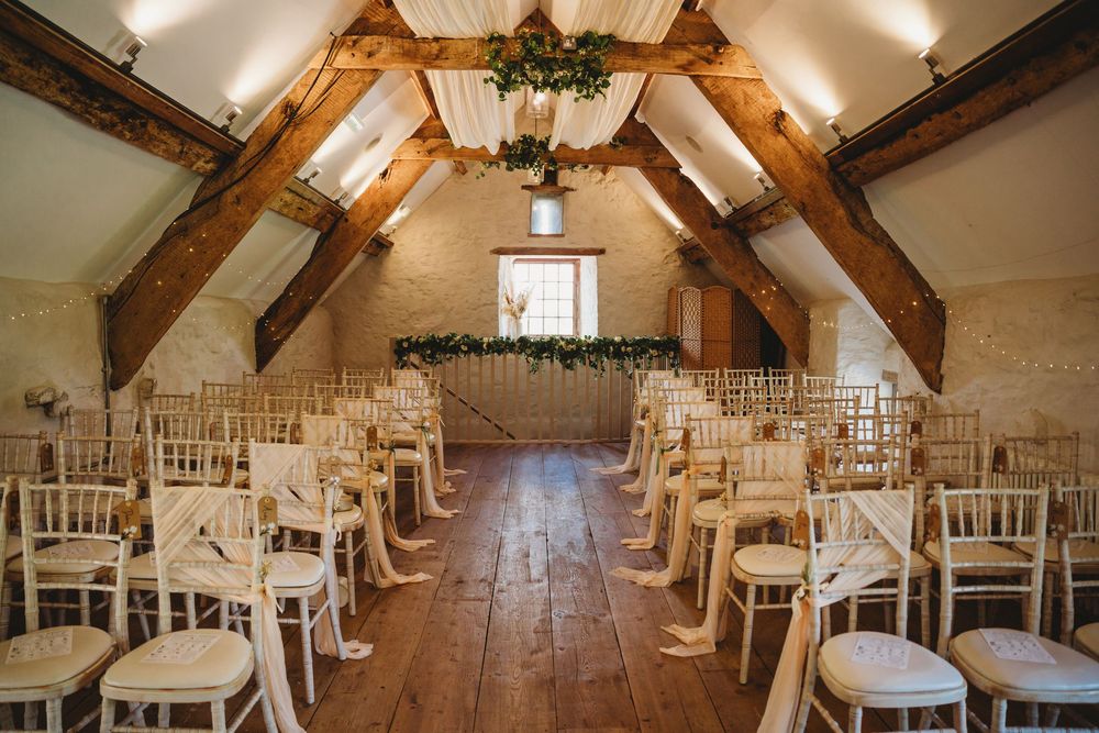 Rustic barn wedding ceremony space with white chairs lining a wooden aisle beneath exposed oak beams and a floral garland.