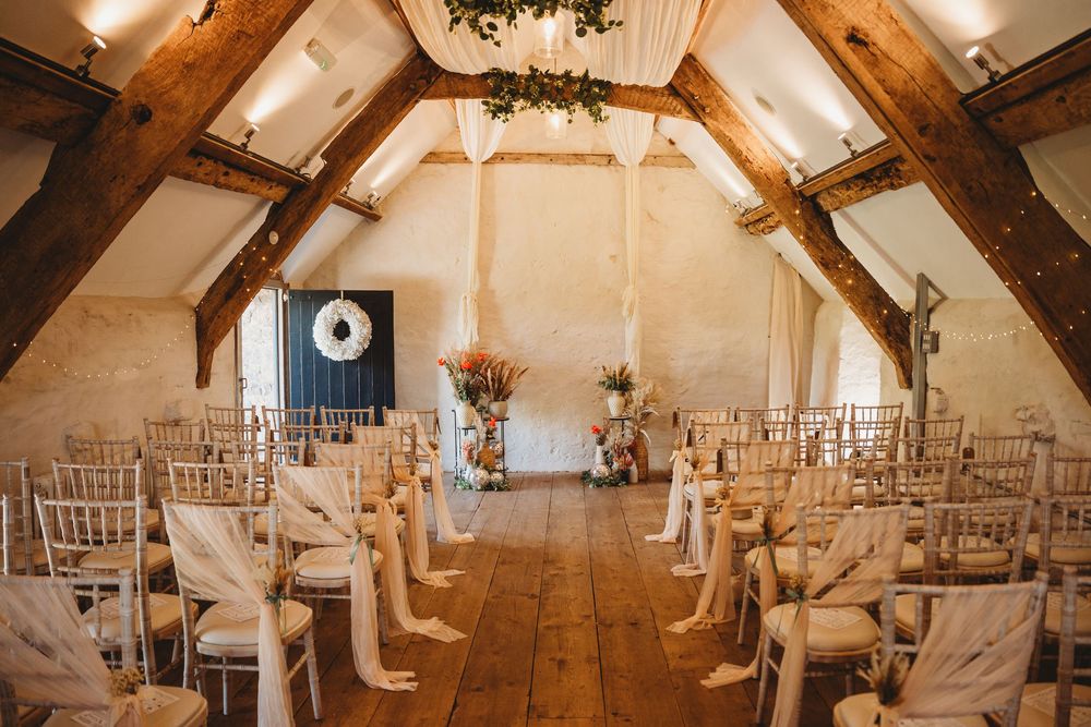 Rustic barn wedding ceremony venue with wooden beams, white chairs with ribbon sashes, floral arrangements and a wreath along the aisle.