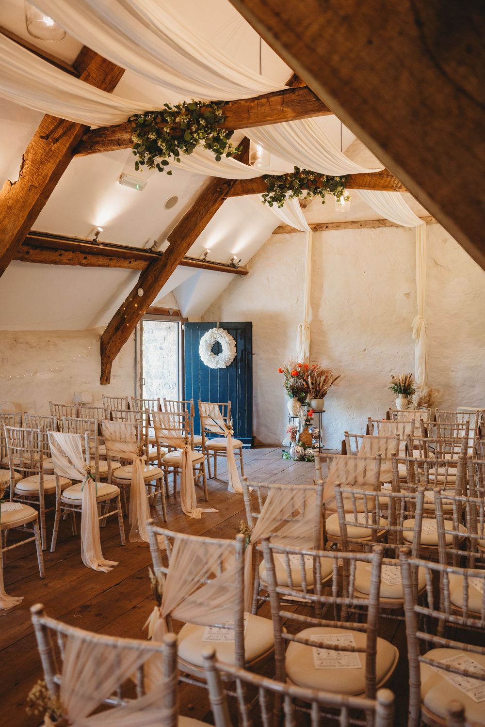 Rustic barn wedding ceremony space with wooden beams, draped fabric, greenery garlands, and rows of chairs with sashes.