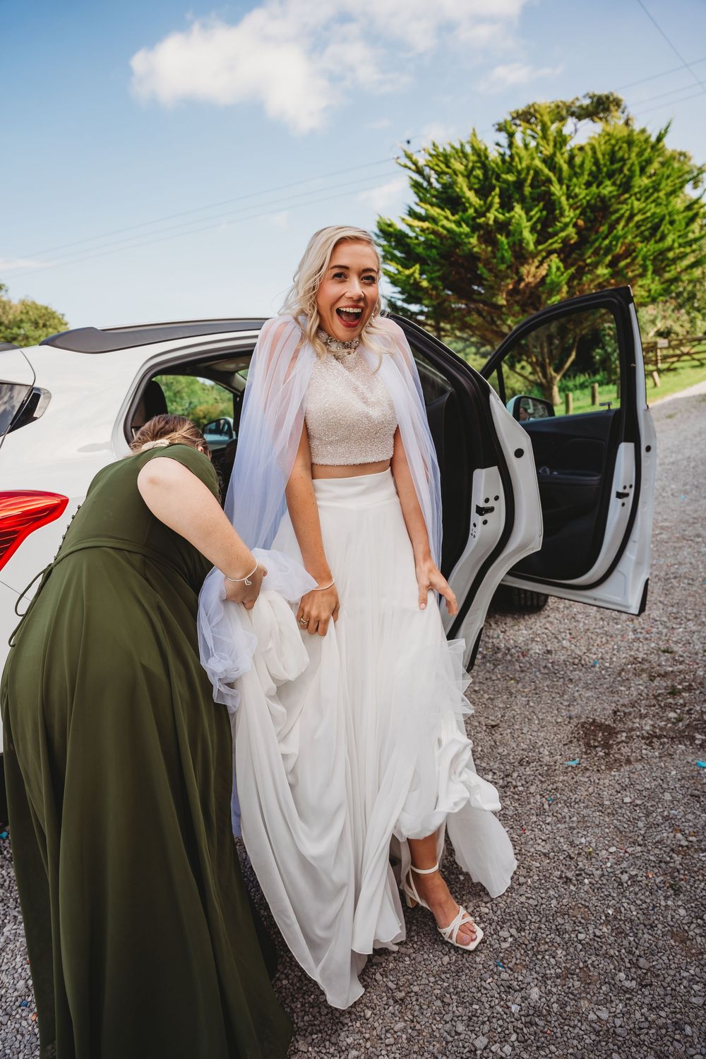 A joyful bride in a white two-piece gown and veil steps out of a white car as a bridesmaid in green helps with her skirt.