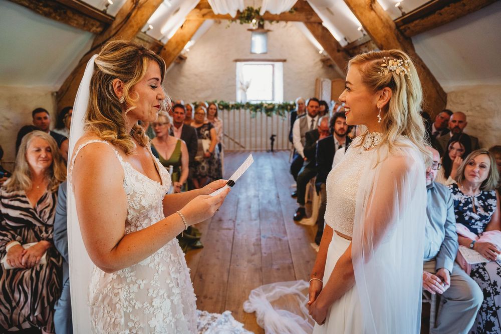 Two brides in white wedding dresses hold hands and read vows during a ceremony in a rustic barn with wooden beams and seated guests.