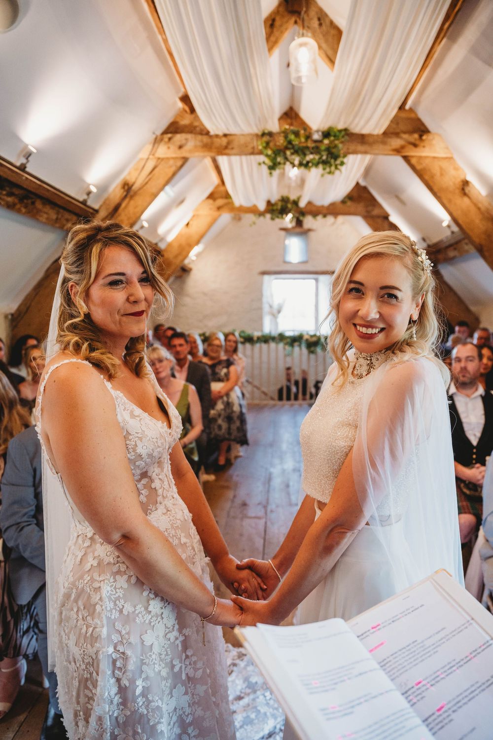 Two smiling brides in lace and tulle wedding dresses hold hands during a ceremony in a rustic barn with exposed wooden beams and guests.