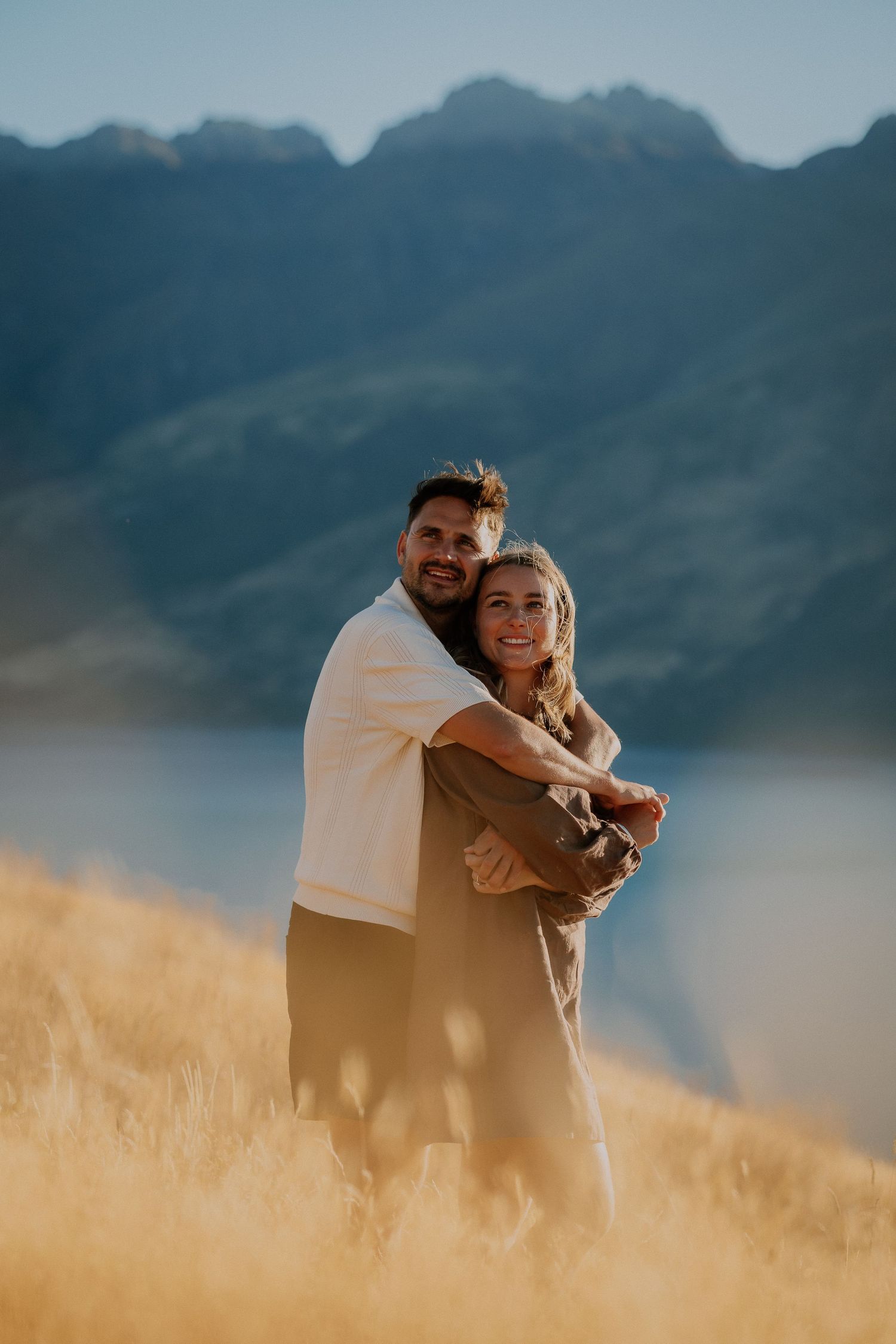 Smiling couple embracing in golden grass on a hillside, with a calm lake and rugged mountains glowing in warm sunset light behind them.
