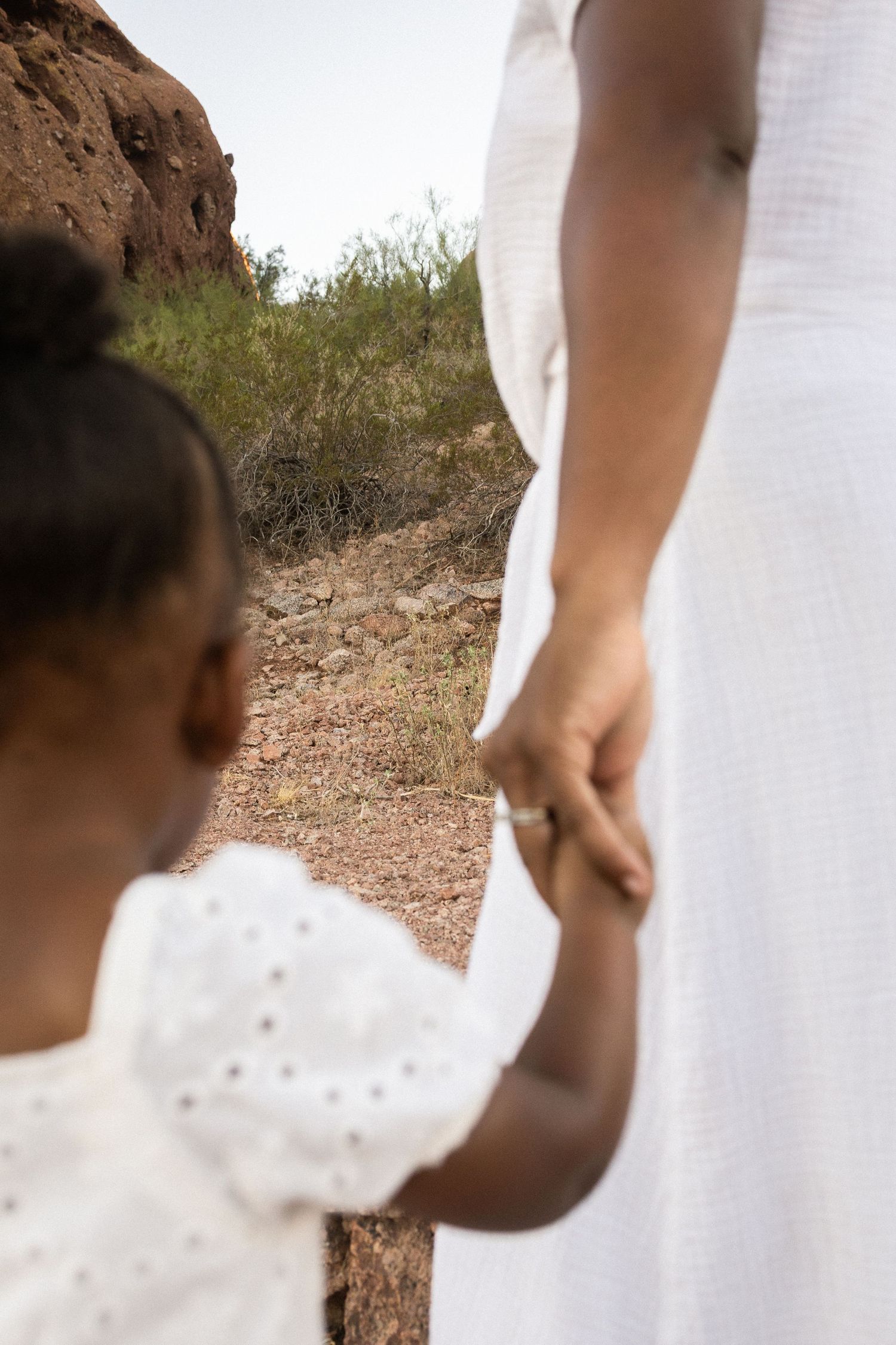 A young girl in white dress holds hands with a woman in white dress on a desert trail at Papago Park in Phoenix.