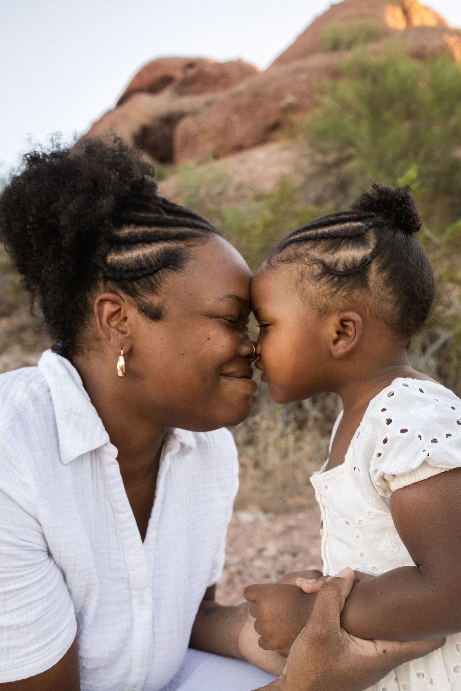 A Black mother and young daughter share a tender nose-to-nose moment outdoors, both wearing white, with red rocks behind them at Papago Park
