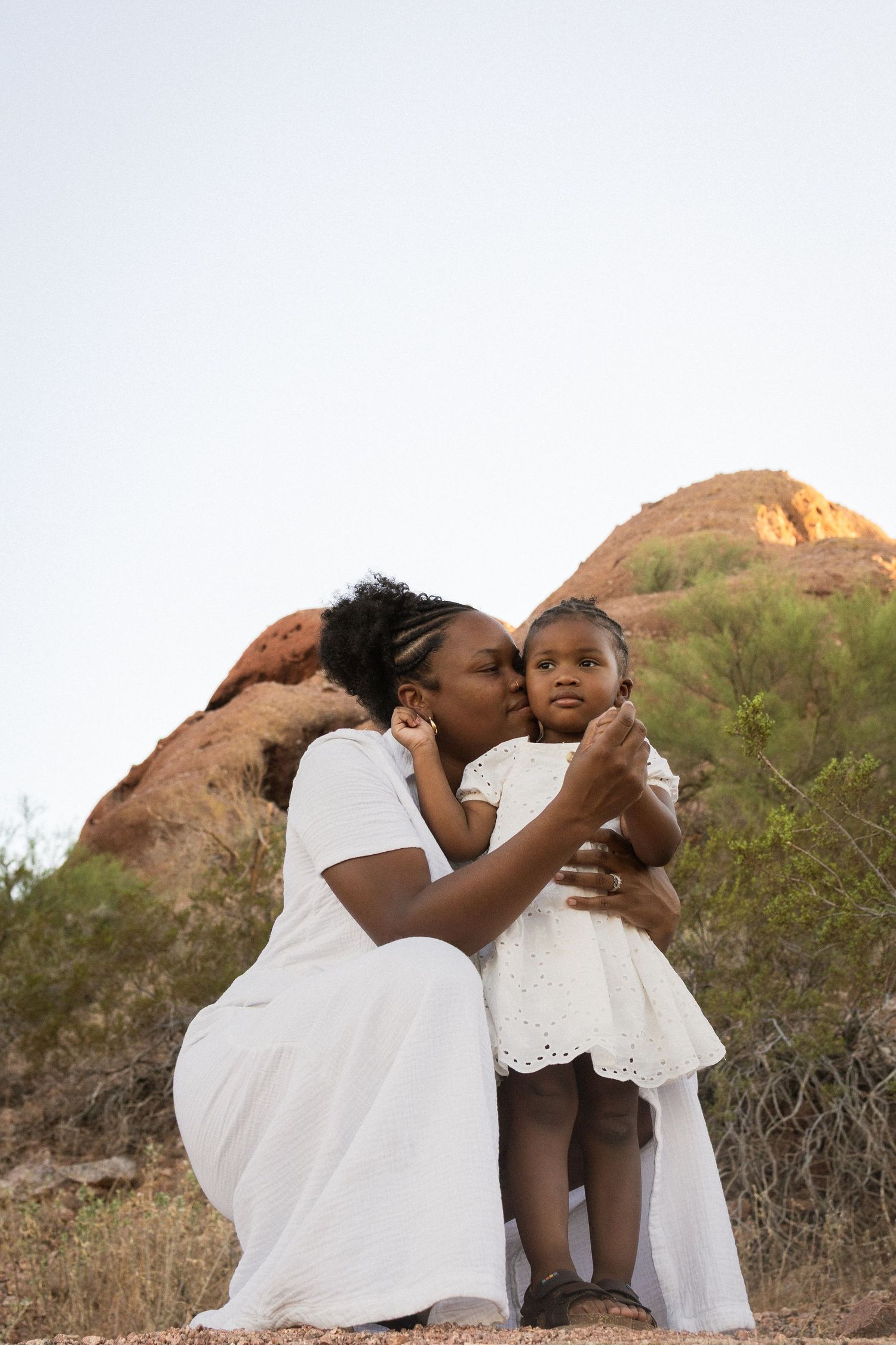 A mother in white dress kisses her toddler daughter outdoors near red rock formations at Papago Park in Phoenix.