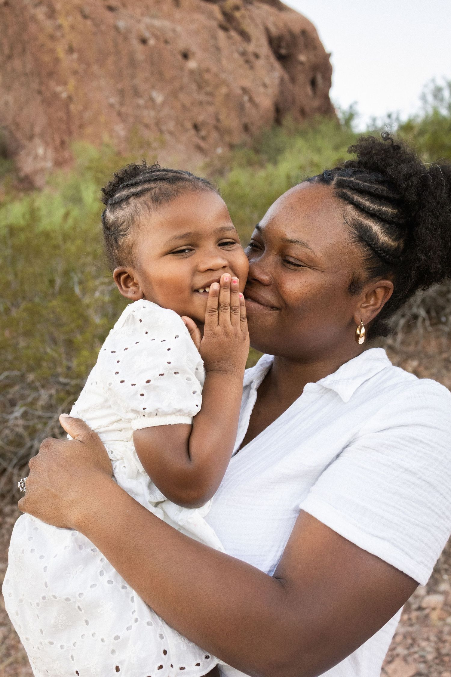 A mother kisses her smiling toddler daughter outdoors, both wearing white, with red rocks and greenery behind them at Papago Park in Phoenix