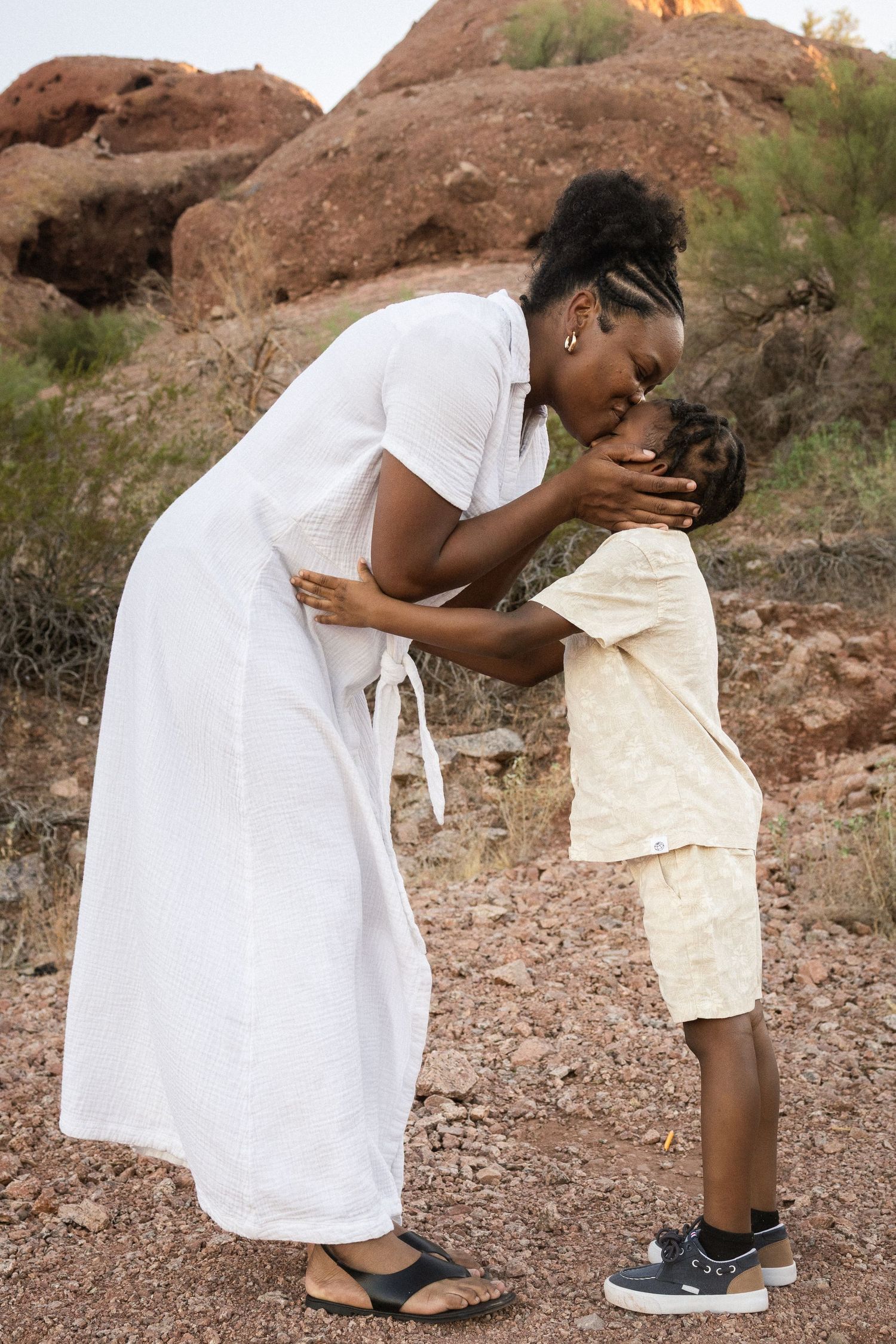A mother in a white dress tenderly kisses her young son outdoors near red rock formations at Papago Park in Phoenix.
