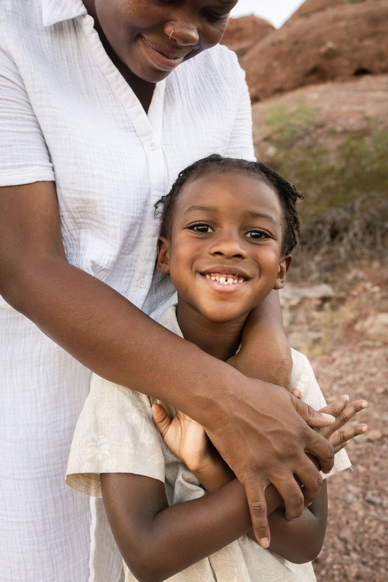 A smiling Black child is warmly embraced by a woman in a white shirt outdoors near red rock formations at Papago Park in Phoenix.