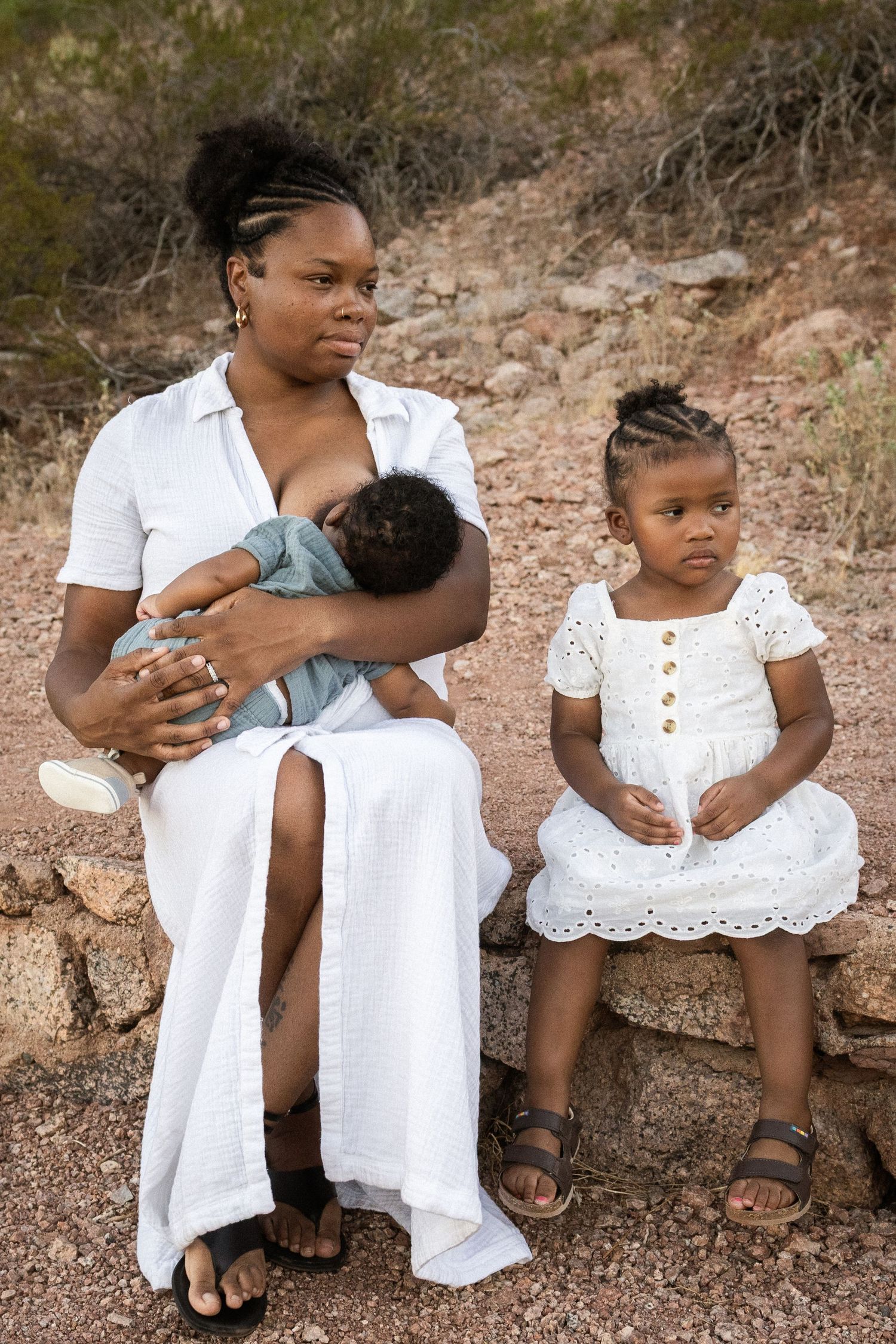 A mother in white dress breastfeeds newborn while young daughter sits beside her on rocks at Papago Park in Phoenix.