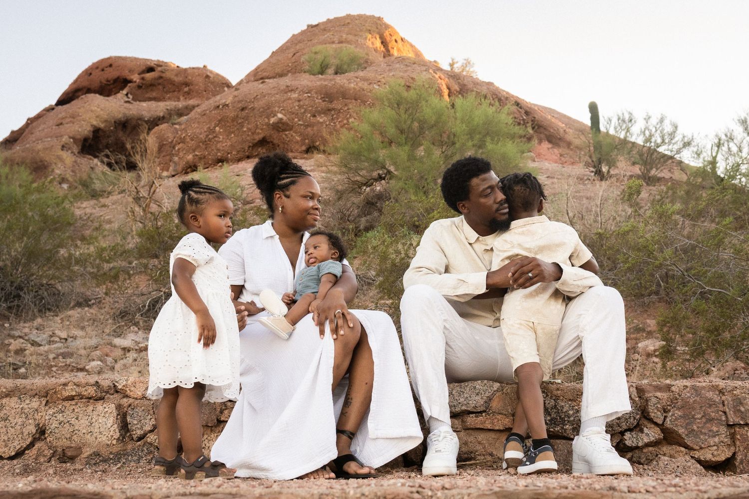 A Black family of four sits outdoors at Papago Park in Phoenix, wearing white and cream outfits, with red rock formations behind them.