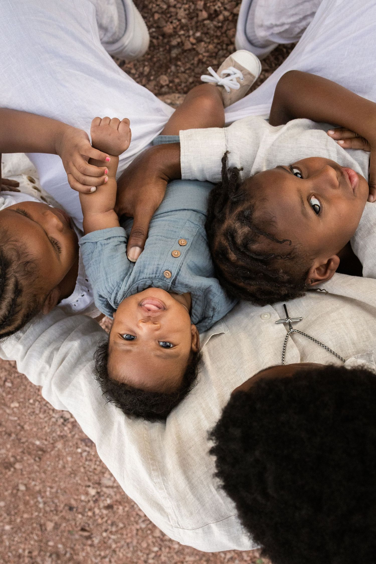 Aerial view of three young Black children lying on a white blanket outdoors, with adults in white clothing beside them at Papago Park.