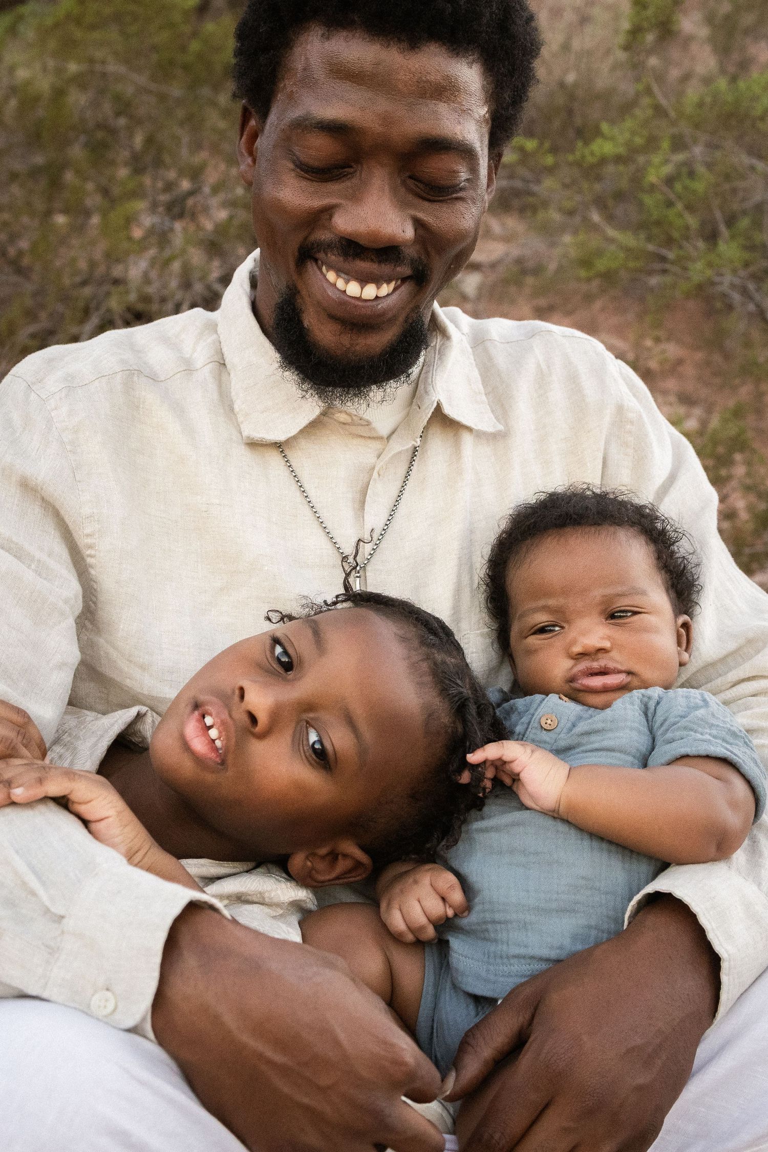 A joyful Black father smiles while holding his two young children on his lap outdoors at Papago Park in Phoenix.