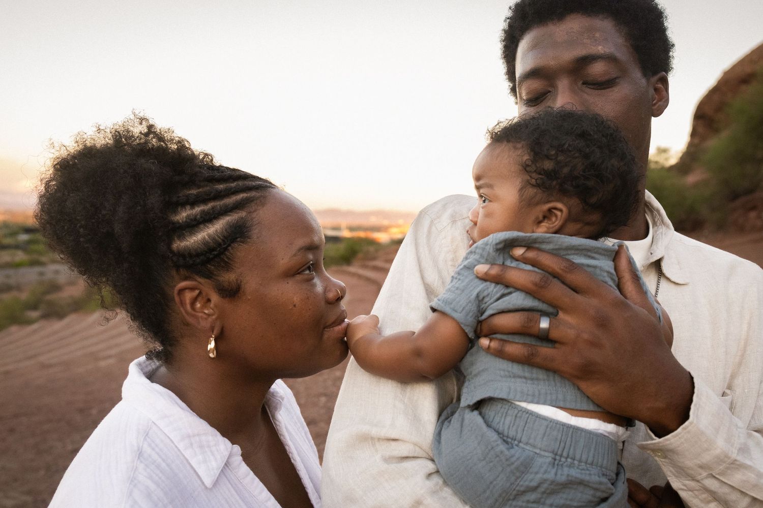Black family with mother kissing newborn baby held by father outdoors at sunset in a tender moment at Papago Park in Phoenix.