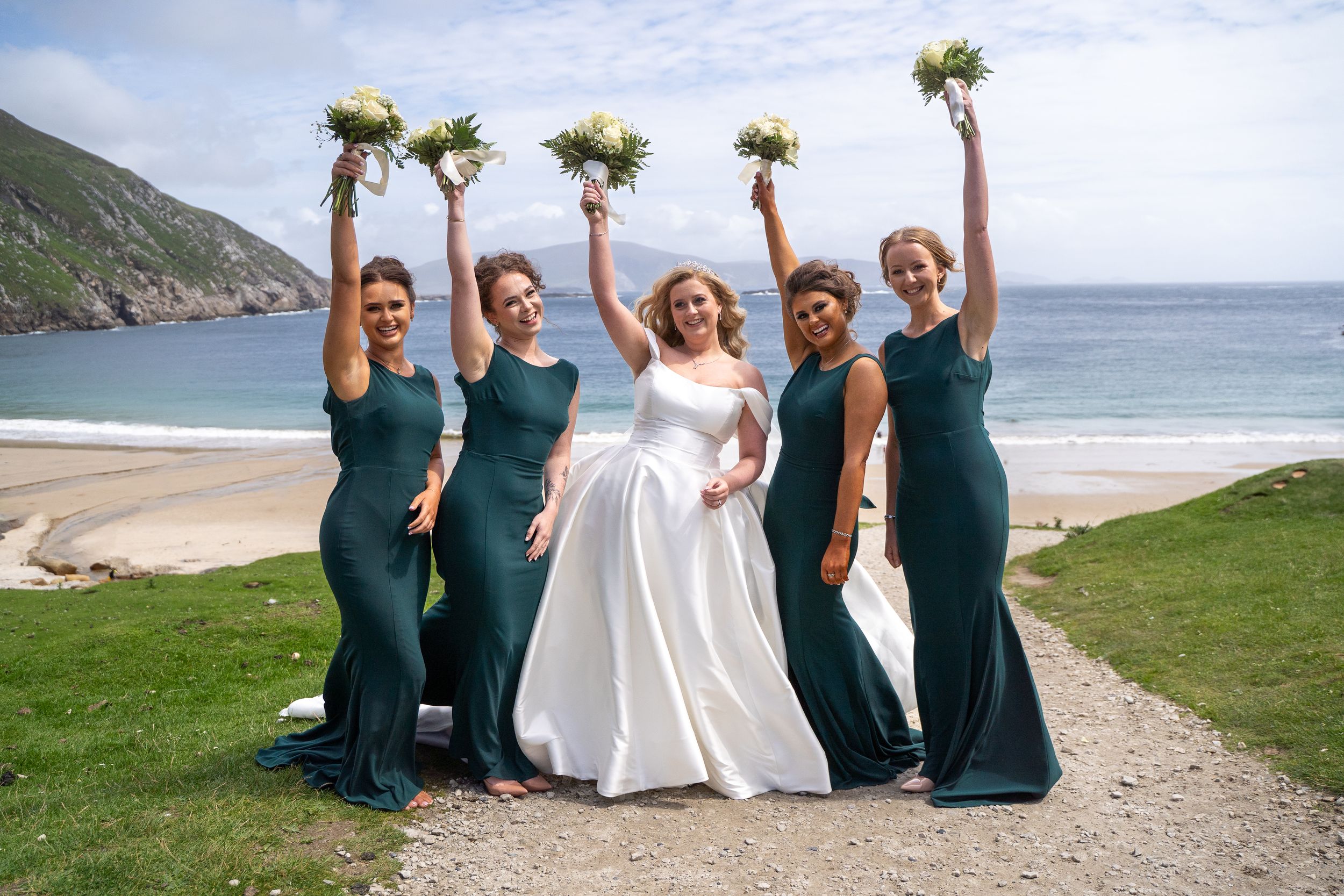 Bride in white gown with four bridesmaids in emerald green dresses raising bouquets on scenic Irish beach.