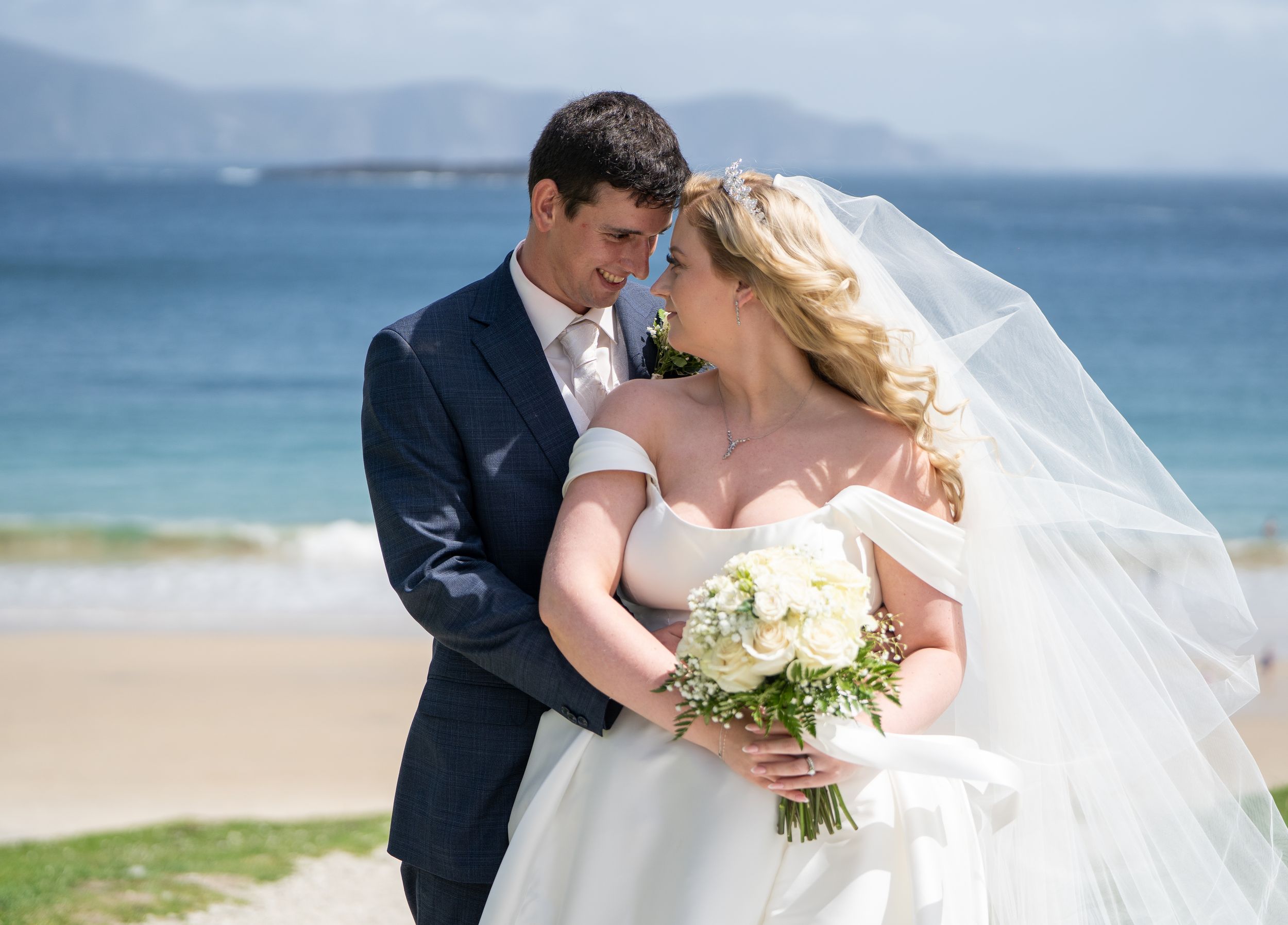 Bride and groom share a romantic moment on a beach, with the bride holding white flowers and her veil flowing in the breeze.