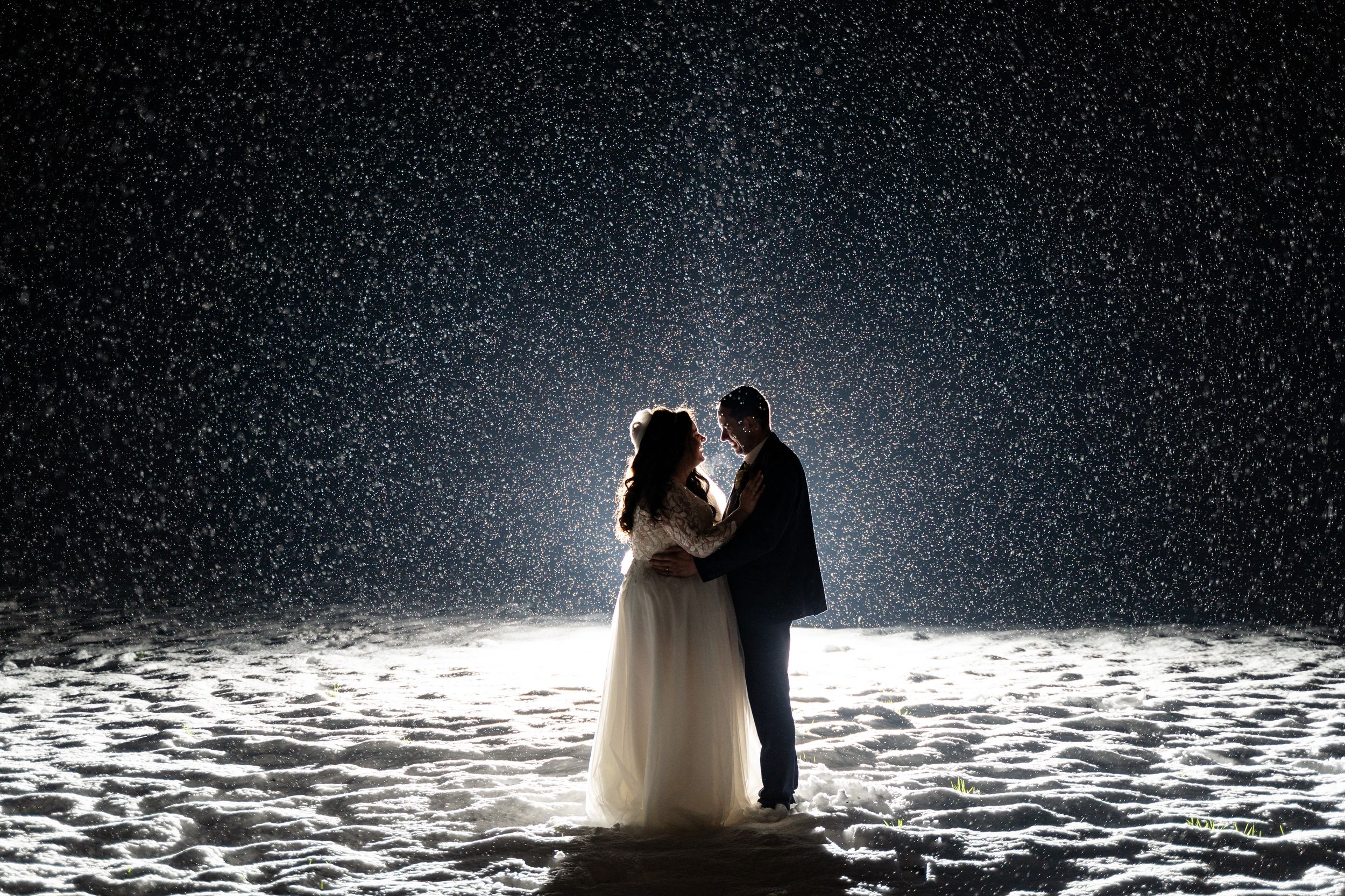 Bride and groom stand together in falling snow at night, illuminated by soft light in a romantic winter wedding scene.