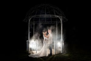 Bride and groom embrace under illuminated gazebo at night with dramatic fog and backlighting in romantic wedding portrait.
