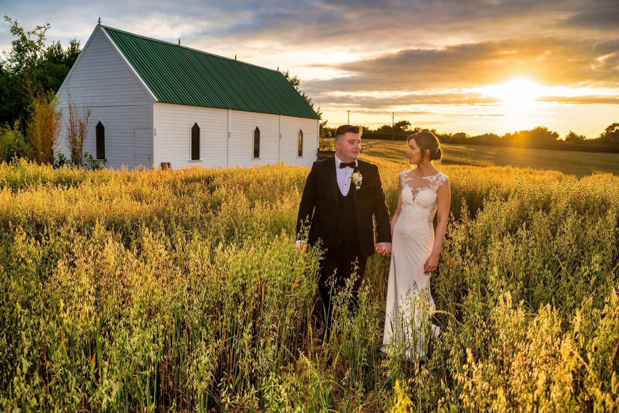 Bride and groom hold hands in a golden wheat field at sunset at the Clanard Court Hotel photo by AK wedding photography and videography.