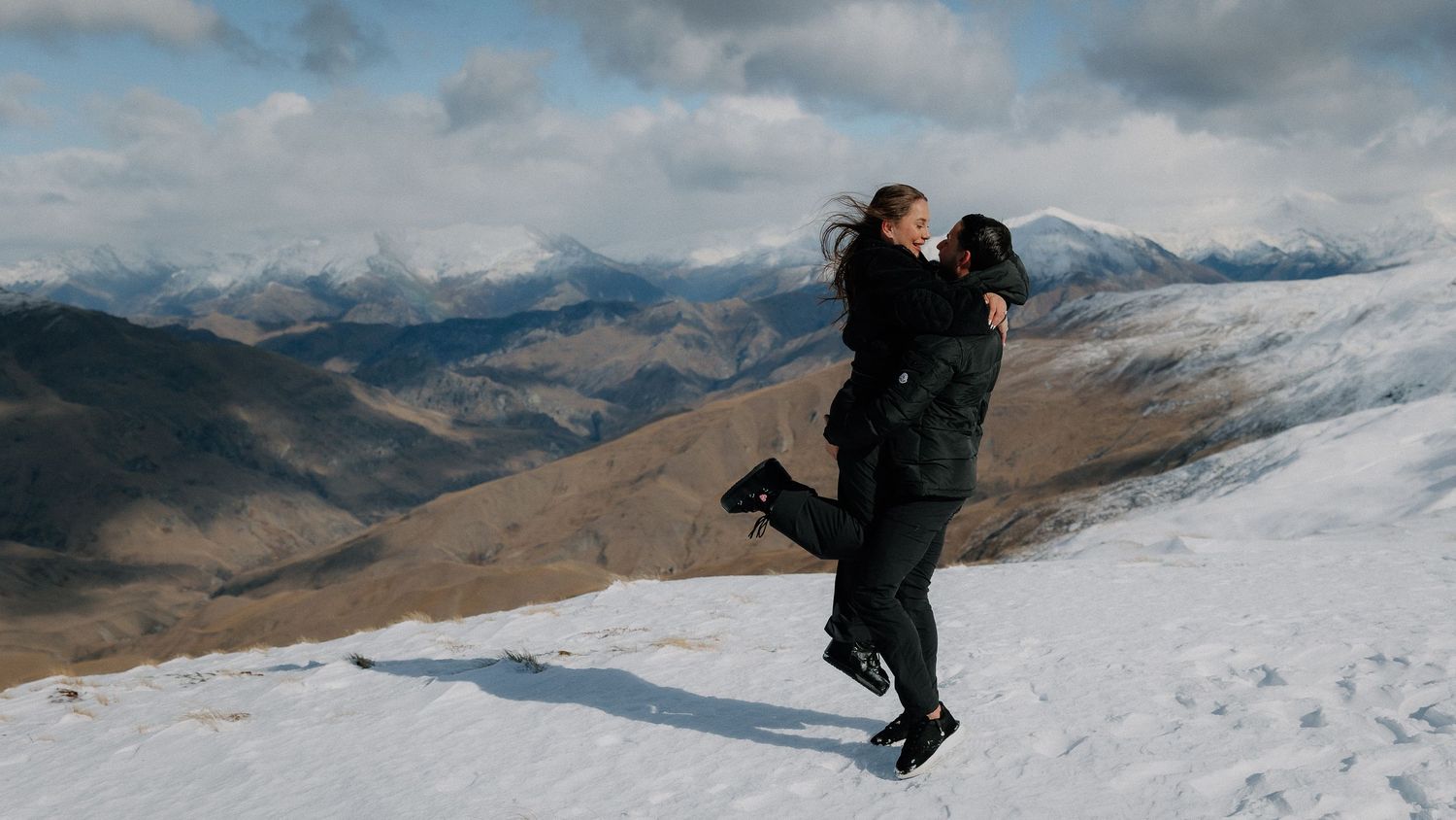 Couple embracing on a snowy mountain slope with scenic winter valley and cloudy sky in background.