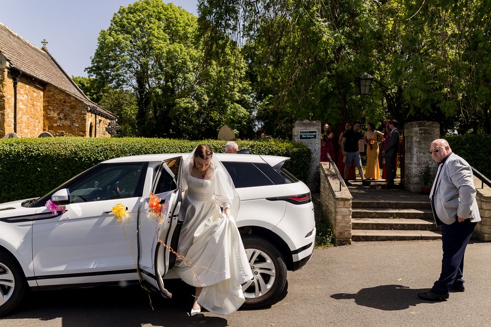 Bride in white gown stepping out of decorated white Range Rover at church wedding venue on sunny day.