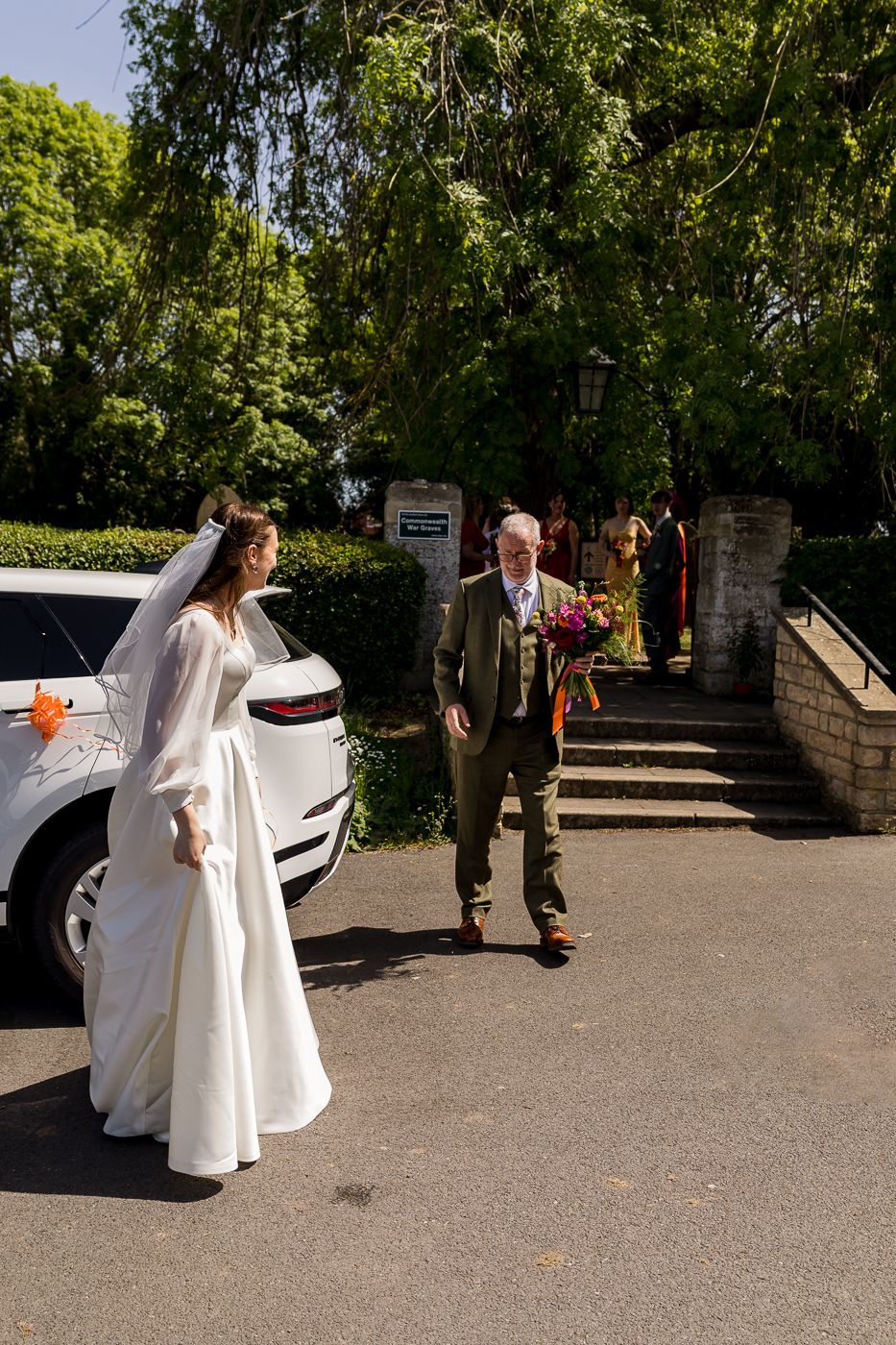 Bride in white gown and veil walks with older man carrying bouquet toward church entrance on sunny wedding day.