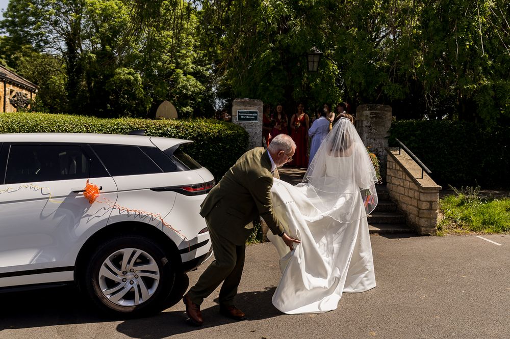 A bride in a white gown and veil arrives at a church in a white SUV as a man helps carry her train.