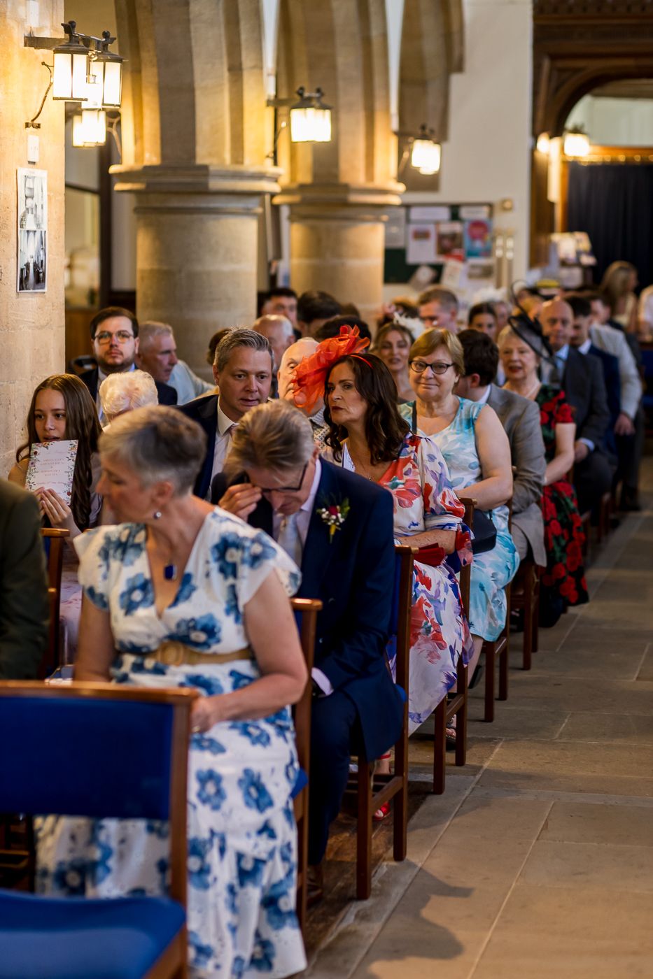 Wedding guests seated in church pews during a ceremony, dressed in formal attire inside a stone church.
