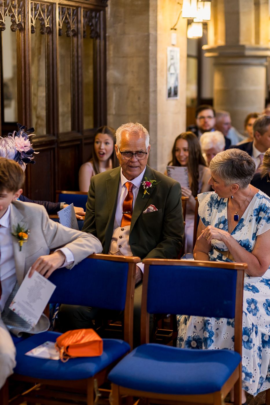 Wedding guests smile and chat in church pews, man in green suit with orange tie stands out among seated attendees.