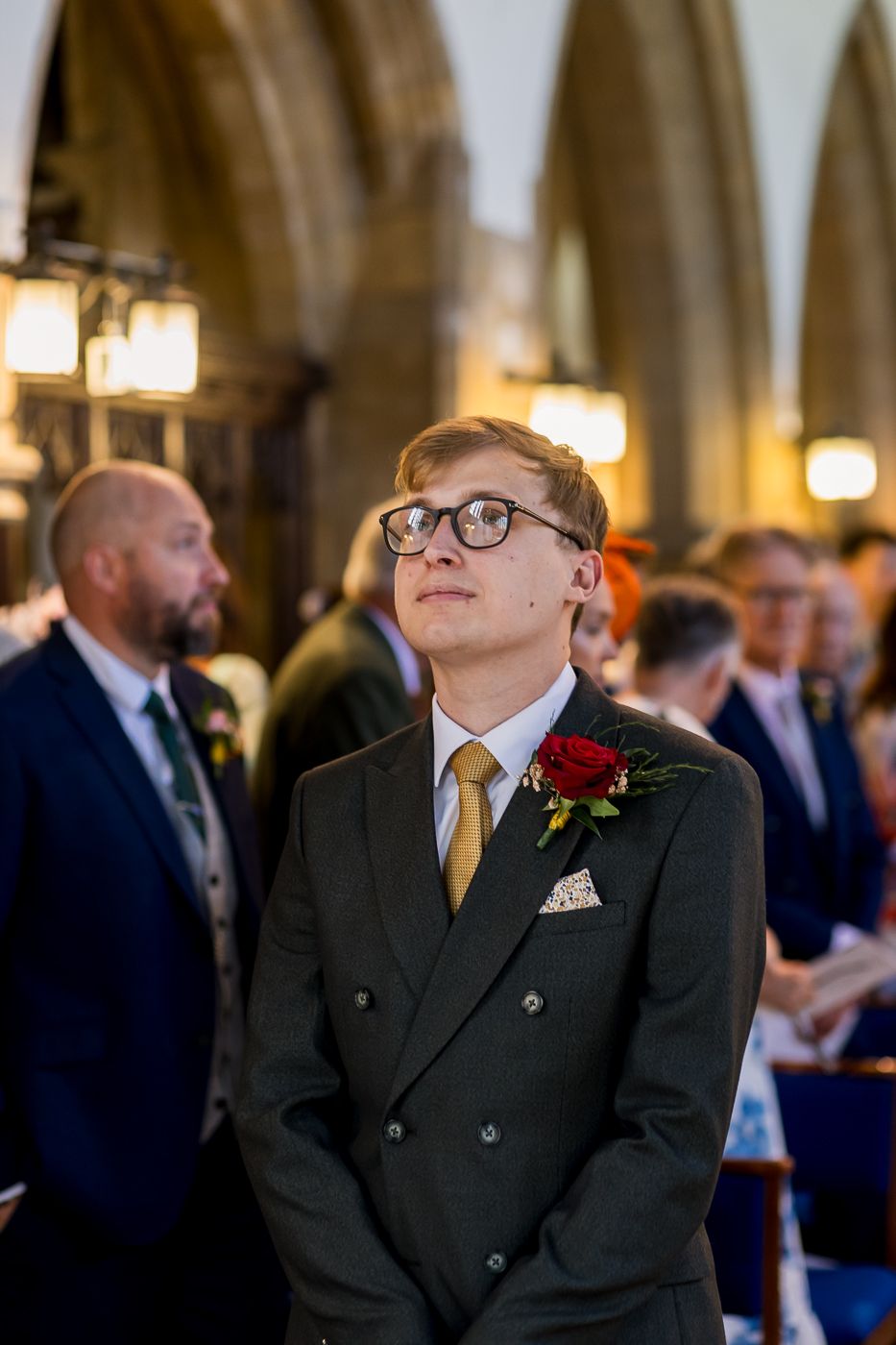 A young groom in a dark double-breasted suit with gold tie and red rose boutonniere looks up during a church wedding ceremony.