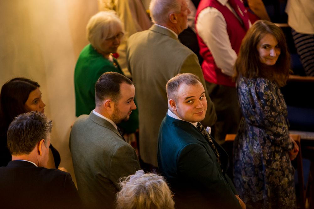 Guests mingling at a wedding reception in a warmly lit venue, with men in suits conversing in the foreground.