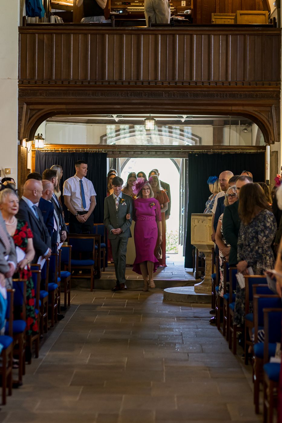 Wedding procession down church aisle with guests standing, wooden gallery above, bride in pink dress walking toward altar.
