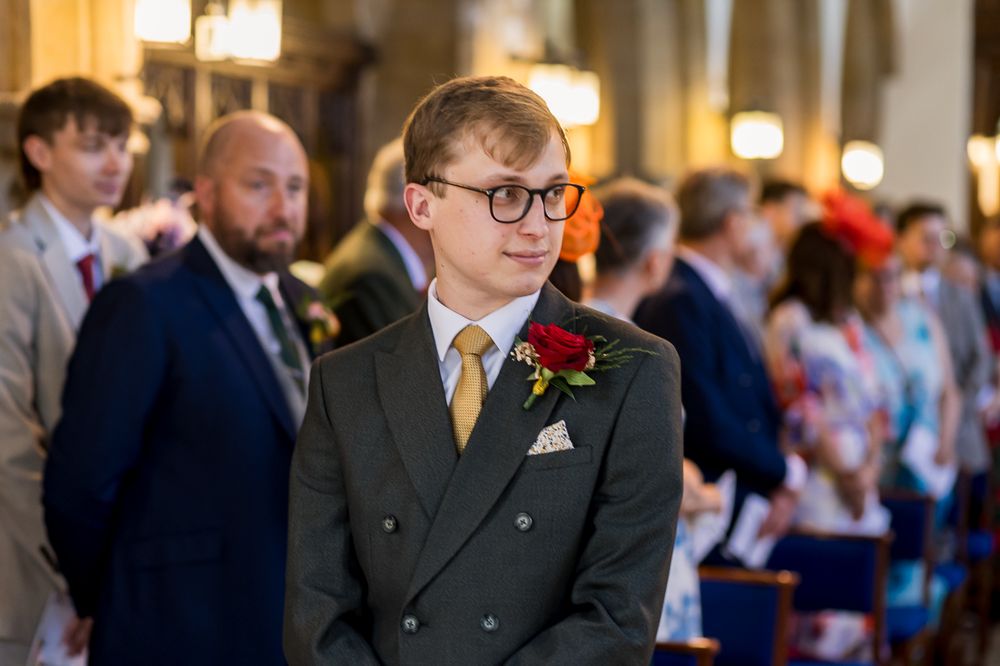 Groom in grey suit with red boutonniere and gold tie awaiting bride at wedding ceremony.