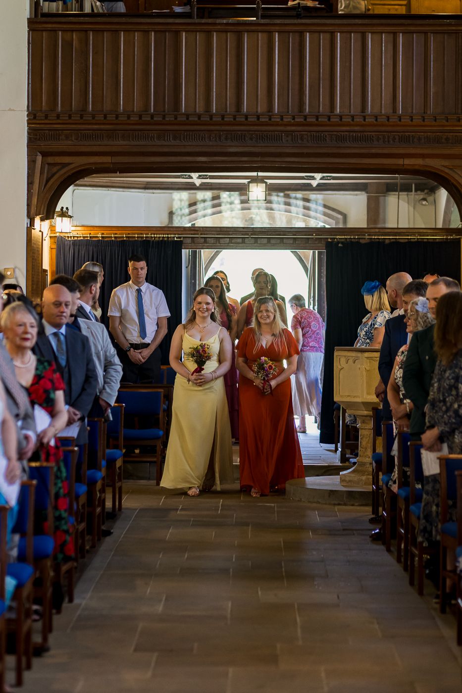 Two bridesmaids in yellow and rust dresses walk down a church aisle during a wedding ceremony.