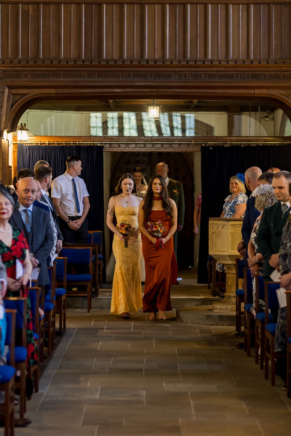 Two bridesmaids in gold and burgundy gowns walk down a church aisle during a wedding ceremony.