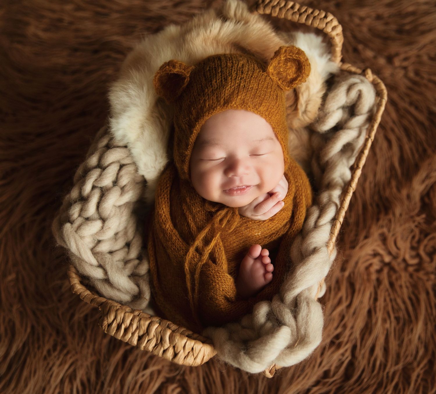 Smiling newborn baby in brown bear knit outfit sleeping in a wicker basket with chunky knit blanket.