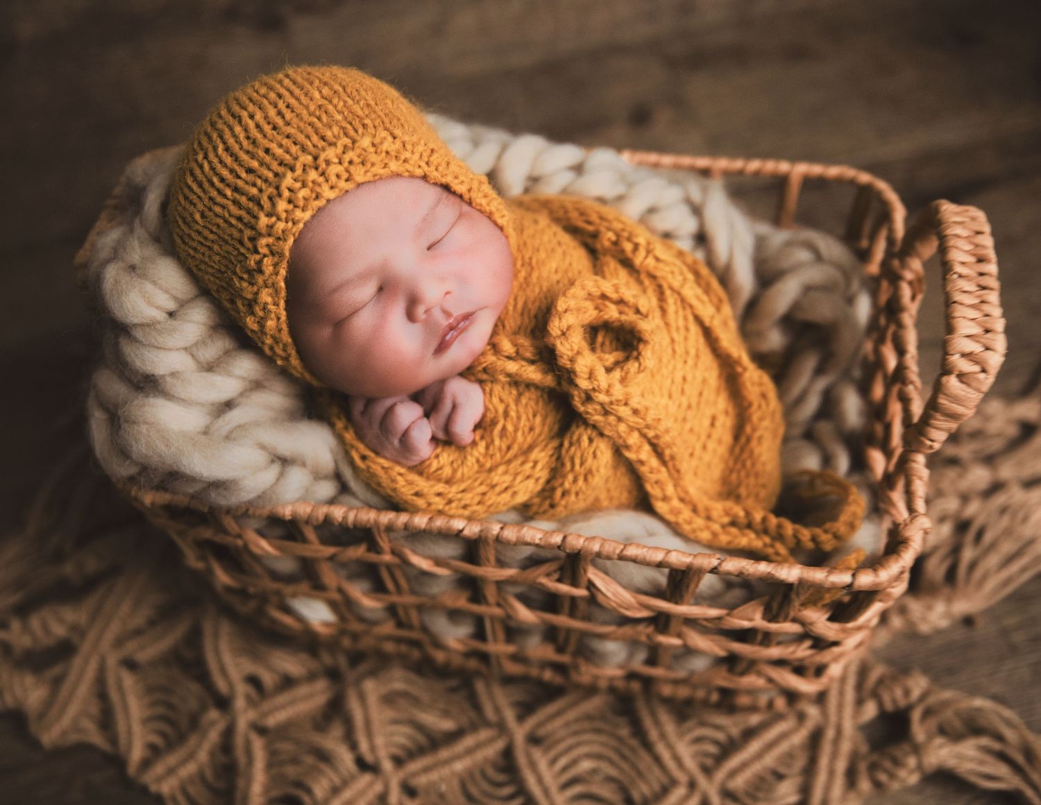 Newborn baby sleeping peacefully in a wicker basket, wrapped in a golden knit blanket and matching bonnet hat.